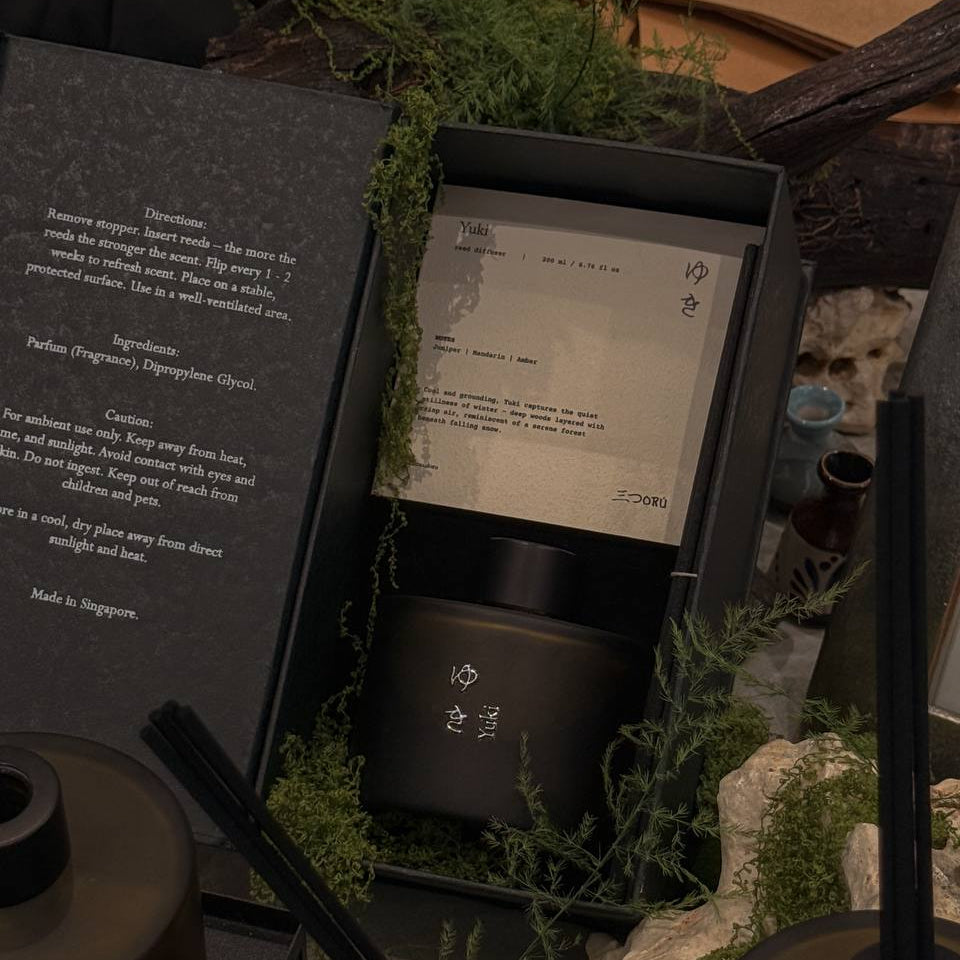 A dark atmospheric product display featuring a black box containing a cylindrical black bottle with white Japanese characters, surrounded by green moss and dried botanicals. An open black card on the left displays product details in English, and other matching black bottles with black straws are partially visible in the foreground.